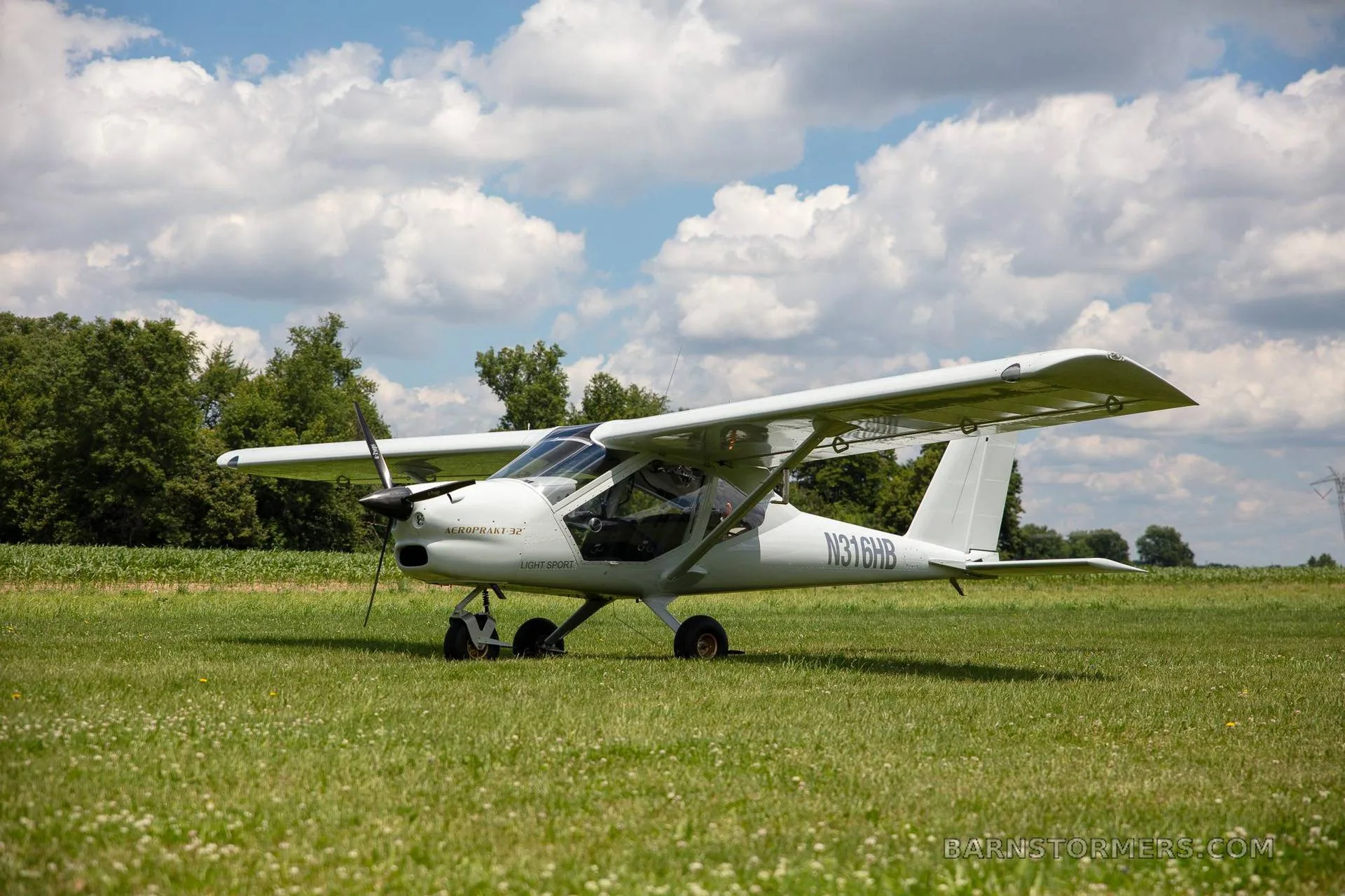 A white Aeroprakt-32 light sport aircraft with registration N316HB sits on a grassy field under a partly cloudy sky.