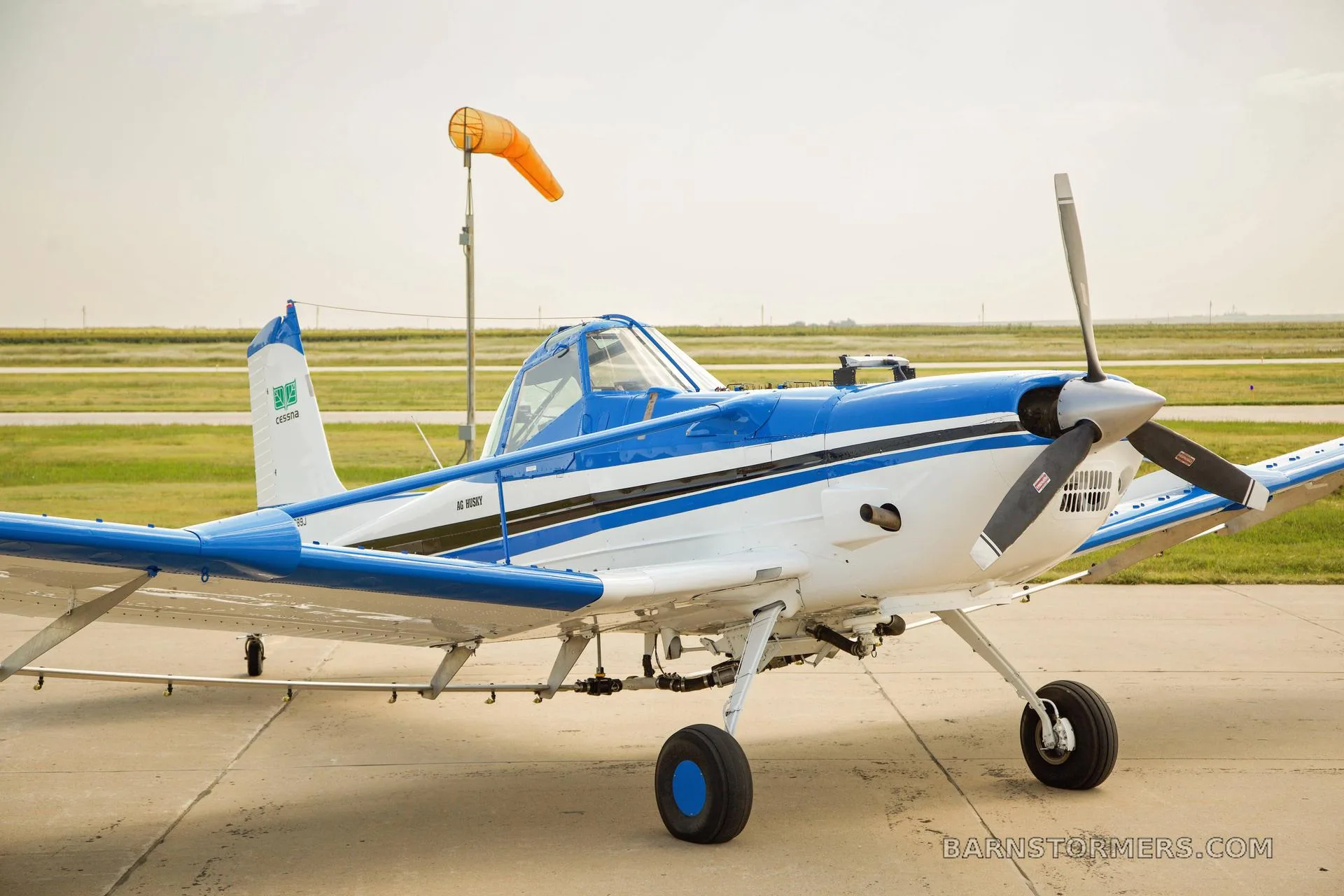 A white and blue agricultural aircraft, possibly a Cessna Ag Husky, is parked on a tarmac with a windsock and grassy field in the background.