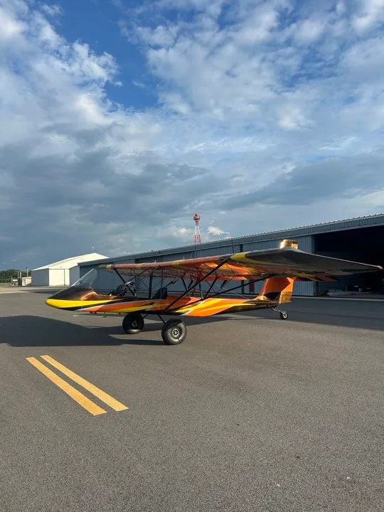 A front-side view of an orange and black ultralight aircraft with a distinctive paint scheme, parked on an asphalt runway near hangars.