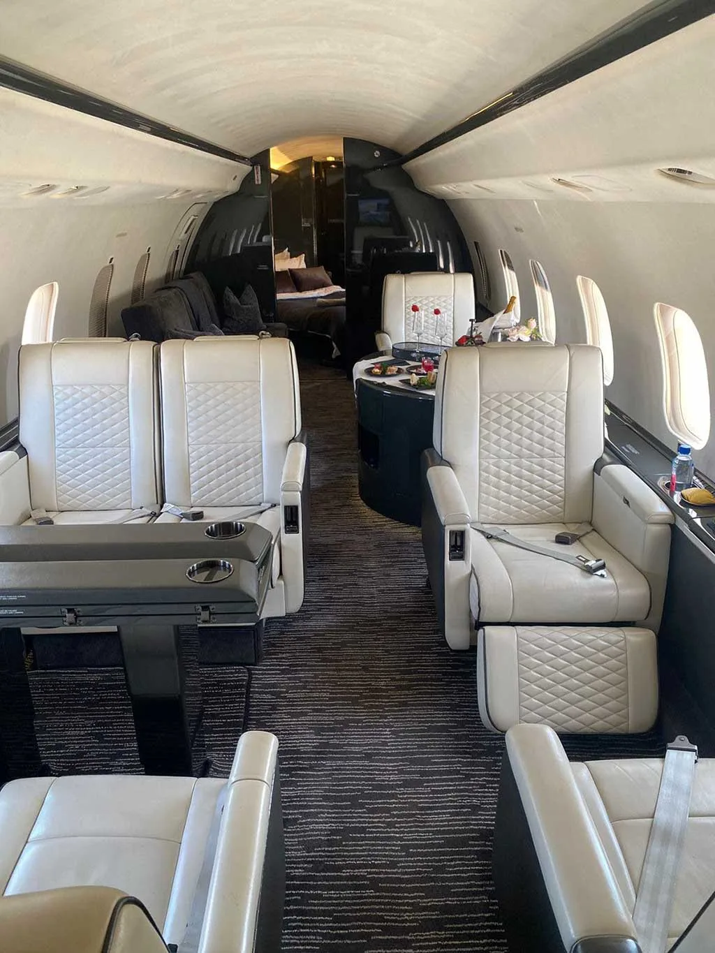 The interior cabin of a private jet with white quilted seats, a dark table set with food and drinks, and a bed visible in the background.