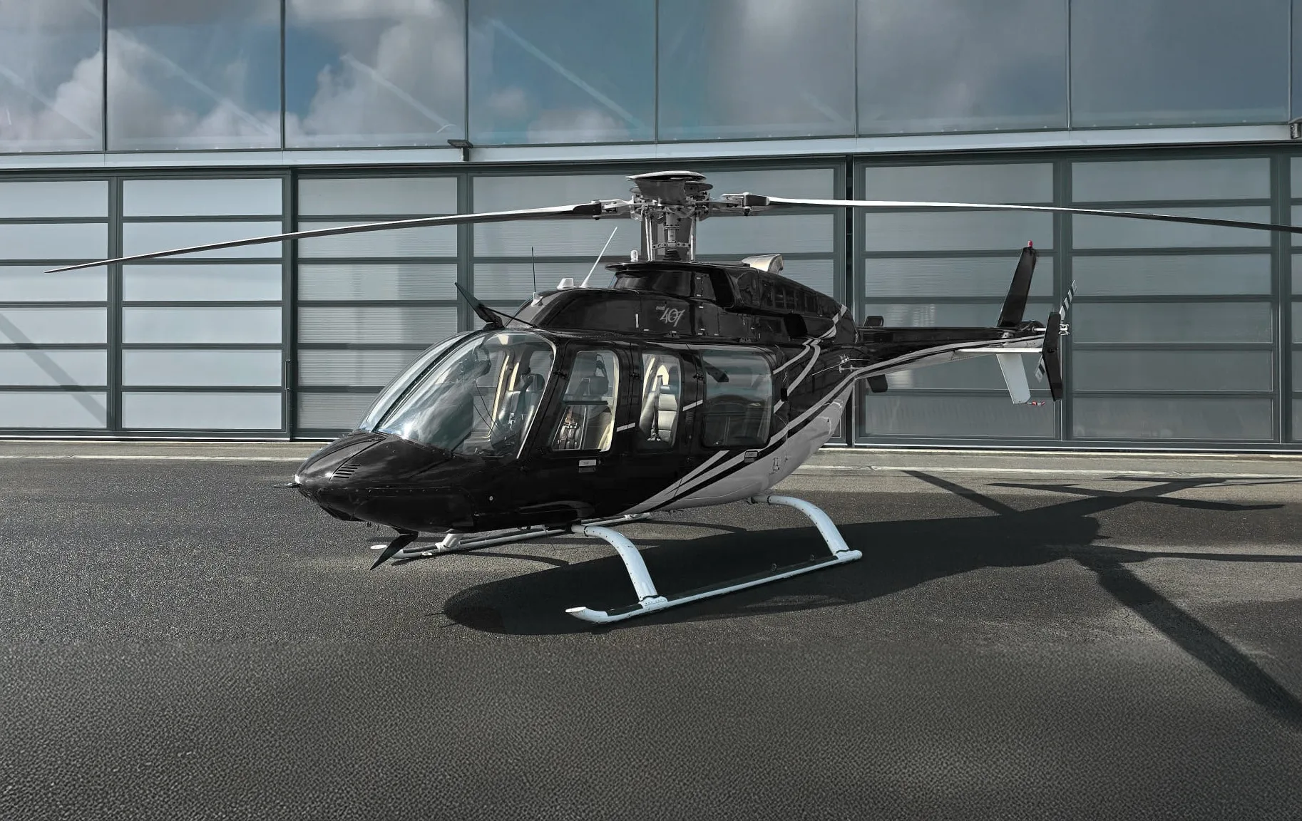 A black and silver helicopter, possibly a Bell 407, parked on an asphalt surface in front of a modern hangar.
