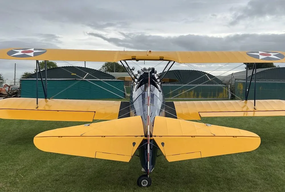 A rear view of a blue and yellow biplane with US military insignia on the wings, parked on a grassy field in front of hangars.