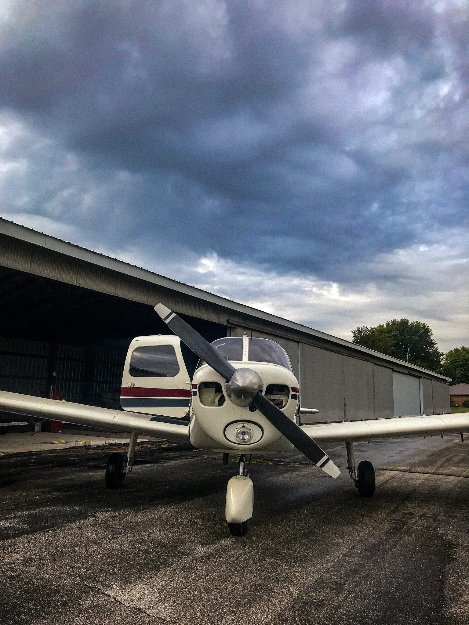 A white single-engine propeller aircraft with red and blue stripes on the tail, parked on an asphalt ramp in front of a hangar under a cloudy sky.