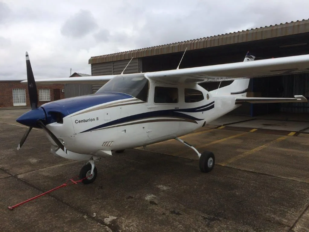 A white Cessna Centurion II with blue and gold stripes is parked on an asphalt apron next to a hangar under a cloudy sky.