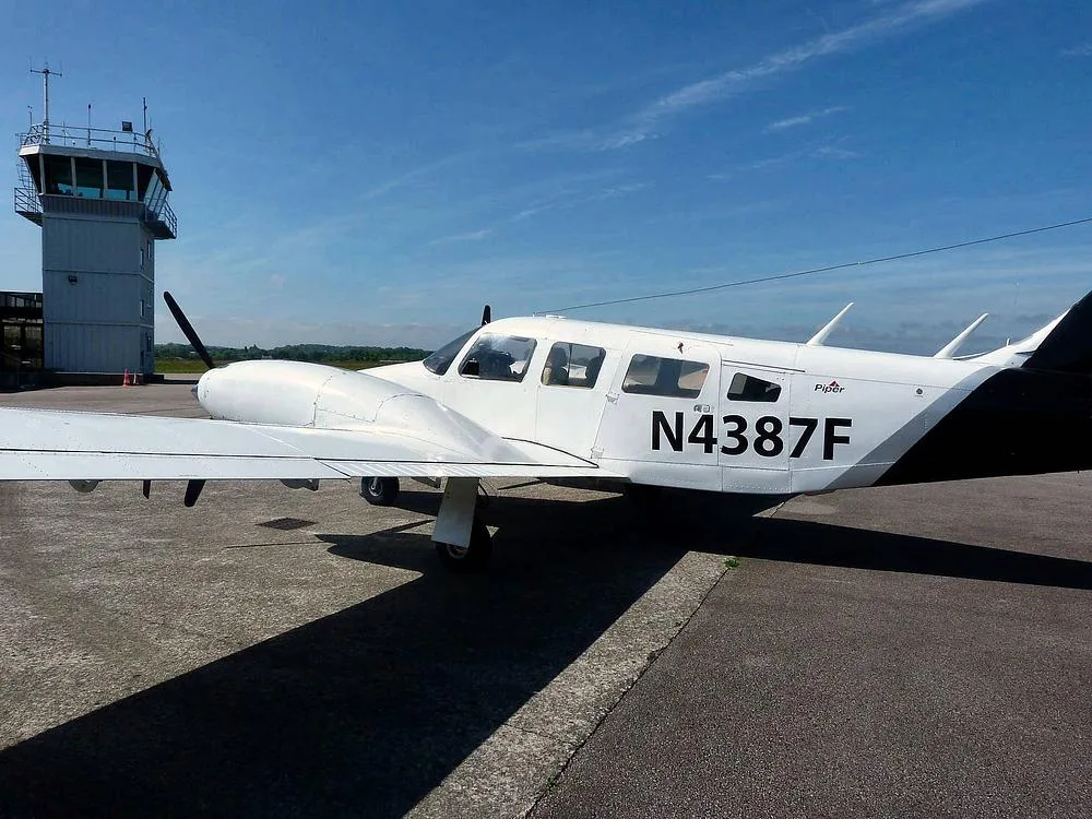 A white and black Piper PA-34 Seneca II, N4387F, parked on an airfield with a control tower in the background under a blue sky.