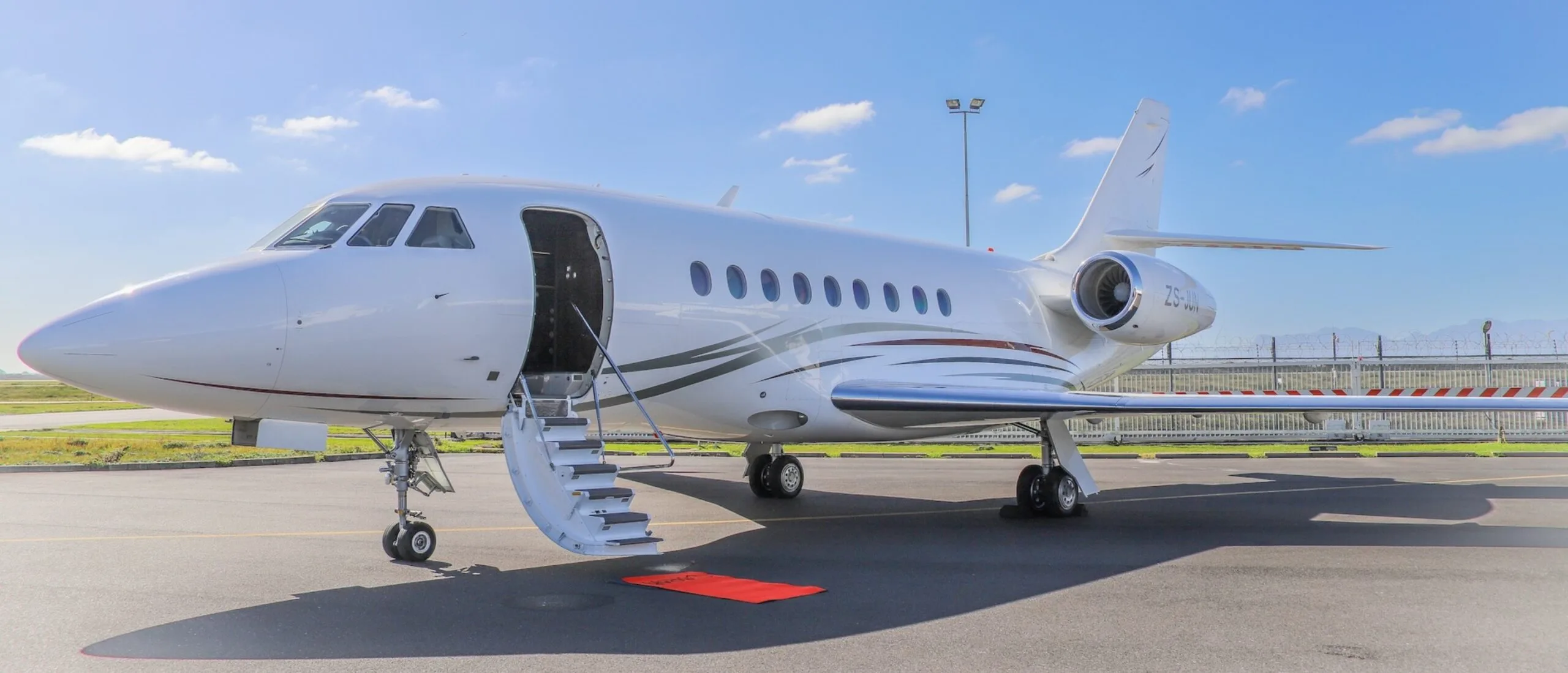A white Dassault Falcon business jet with grey and red stripes is parked on an airport tarmac with its main door open and stairs extended.