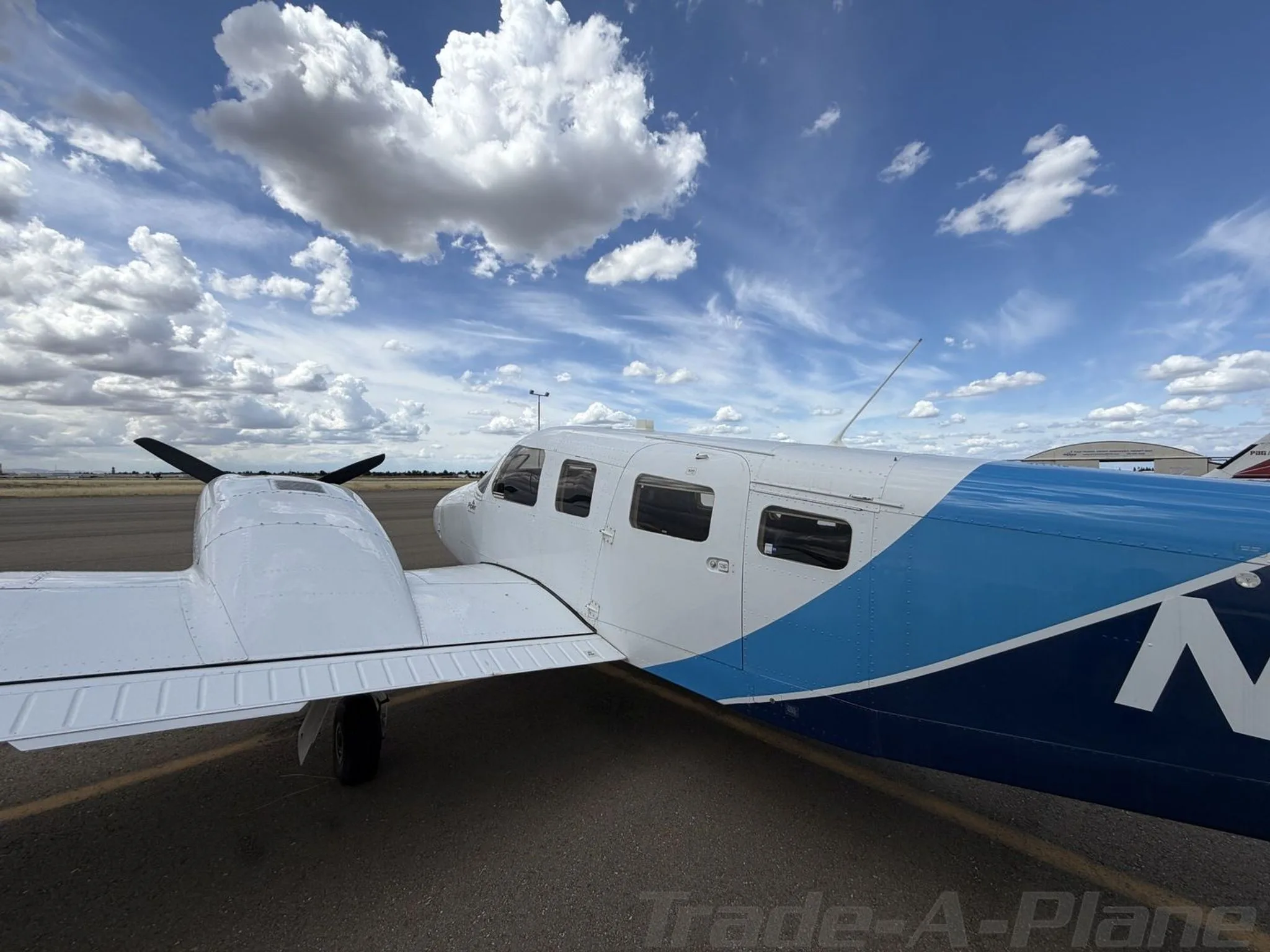 A side-rear view of a white Piper Seneca II aircraft with a blue and light blue paint scheme on the fuselage, parked on an airport tarmac.