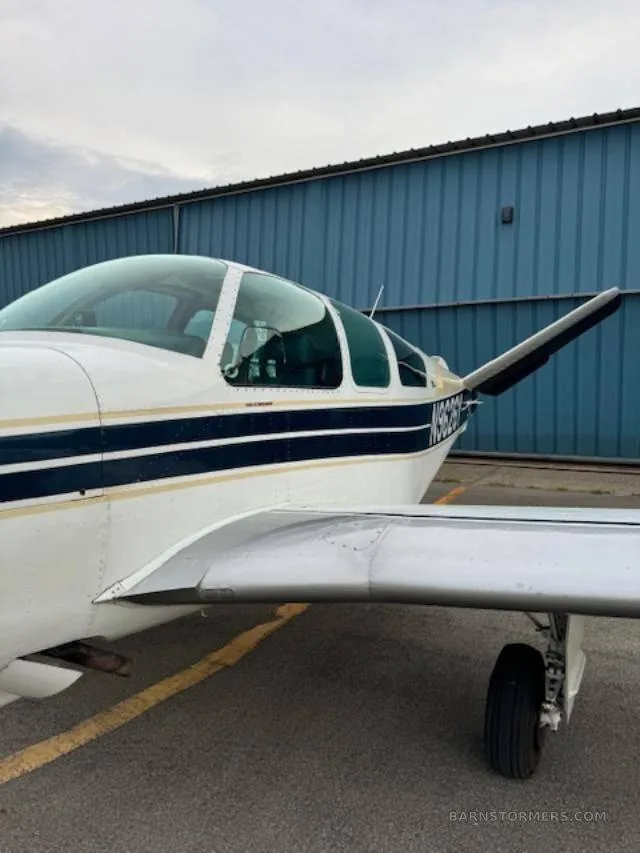 A close-up side view of the cockpit and wing of a white and blue Beechcraft Bonanza, parked in front of a blue hangar.