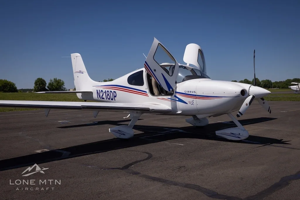 Front-right view of a white Cirrus aircraft (N218DP) with blue and red stripes, doors open, parked on an airfield.