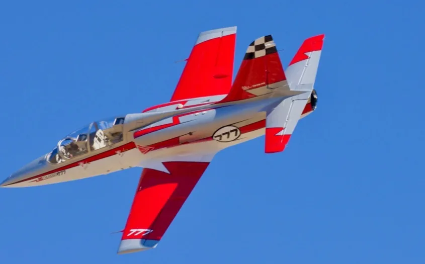 A silver and red jet with "777" markings and a checkered tail flies against a clear blue sky.