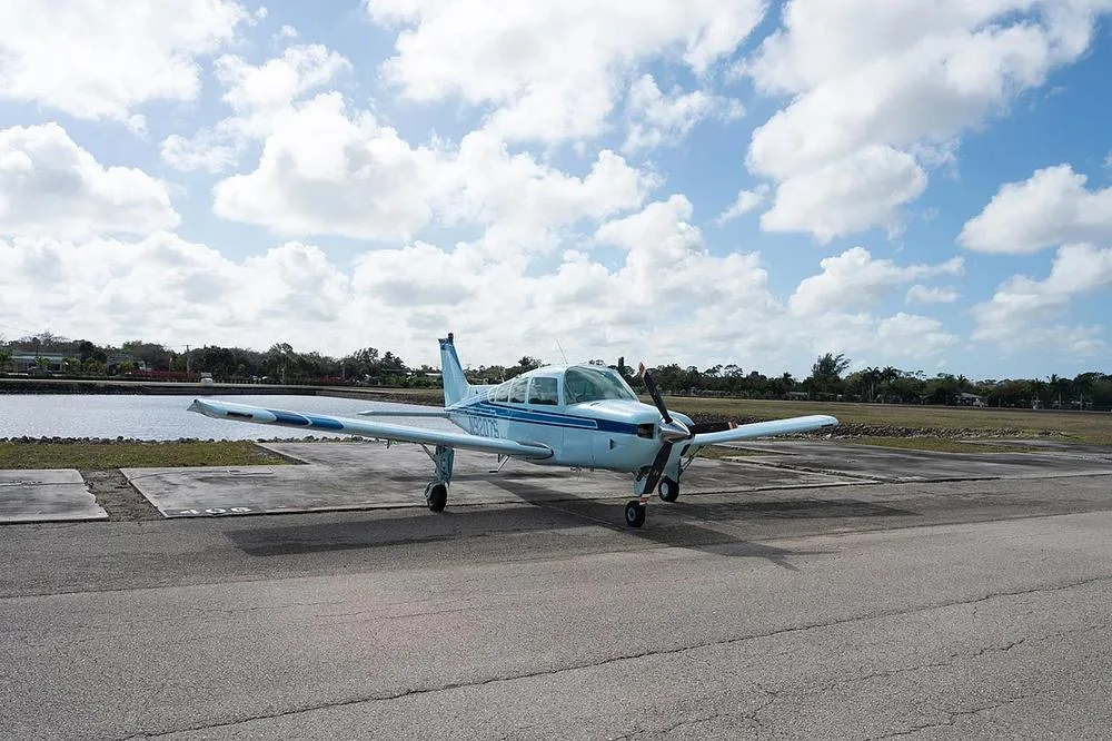 A light blue single-engine propeller aircraft, N9207S, is parked on an asphalt apron, angled towards the viewer, with water and trees in the background.