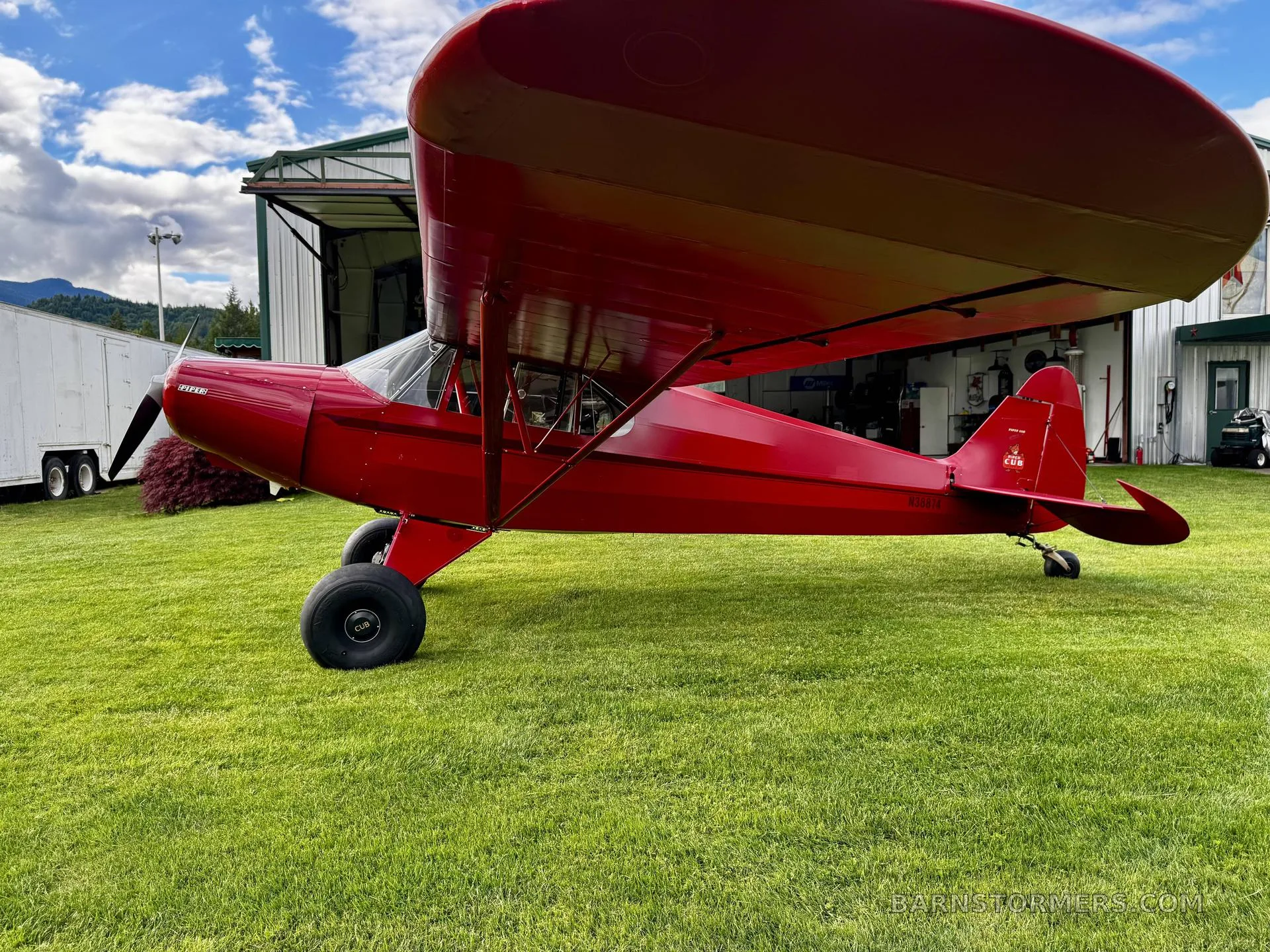 A red Piper Cub aircraft with registration N3081H is parked on green grass in front of a hangar under a partly cloudy sky.