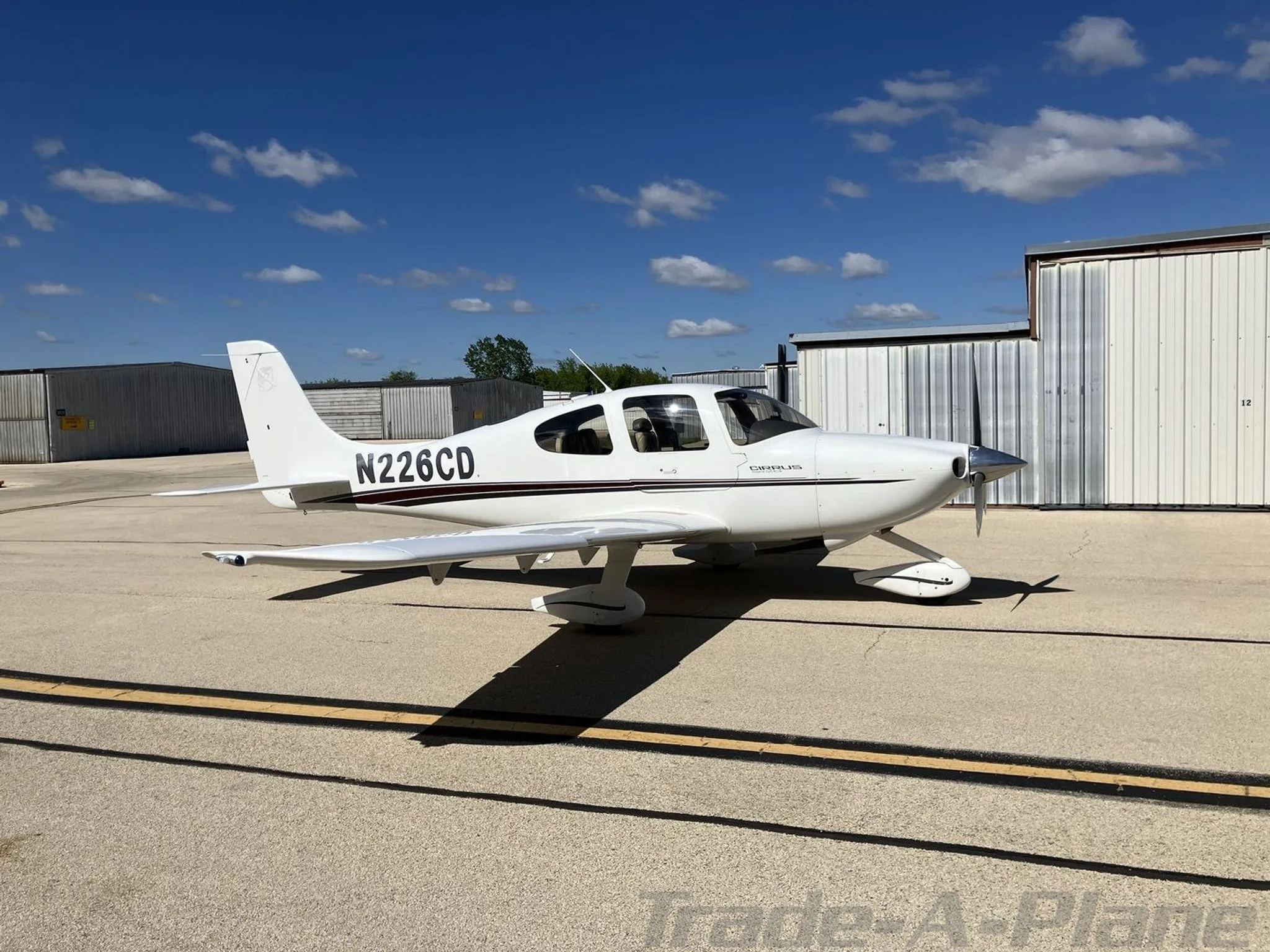 A white Cirrus SR20 aircraft with black and brown stripes, tail number N226CD, parked on an airfield tarmac under a blue sky.