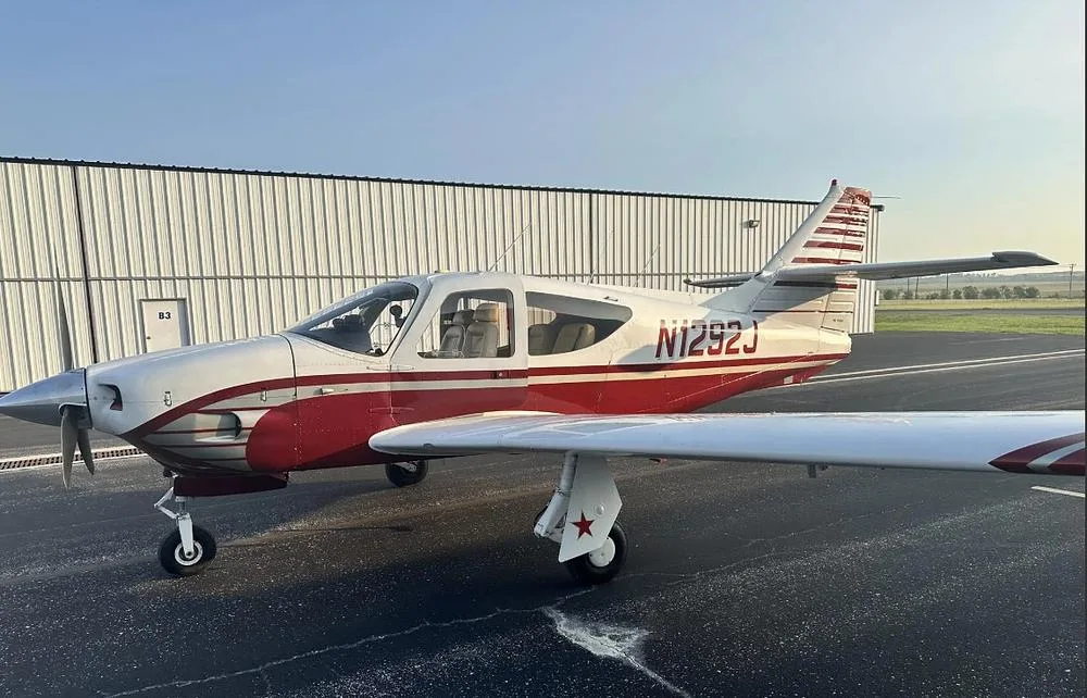 A full side view of the white and red single-engine aircraft N1292J, parked on an airfield next to a hangar.