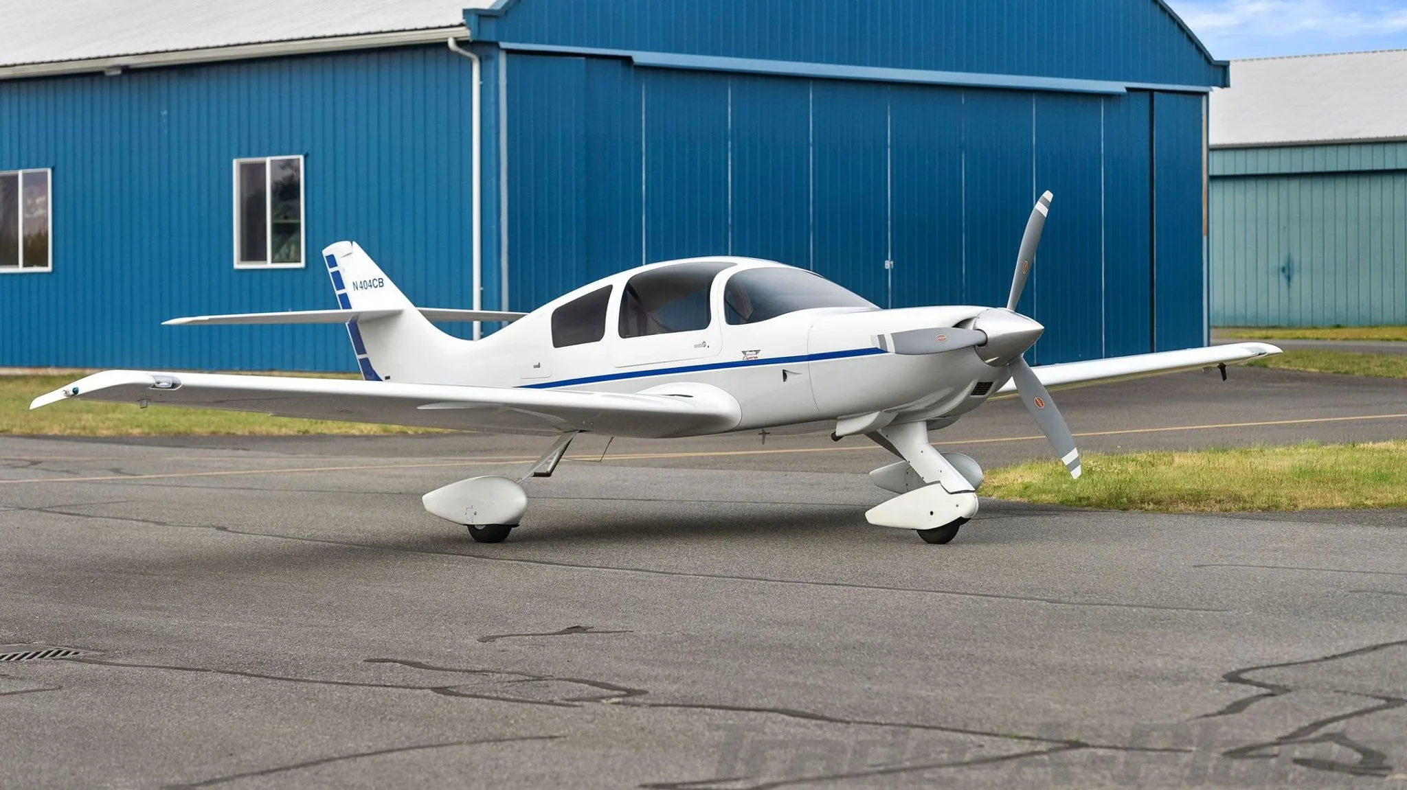 A white single-engine aircraft with blue stripes on the fuselage and tail, registration N404CB, parked on an asphalt runway in front of a blue hangar.