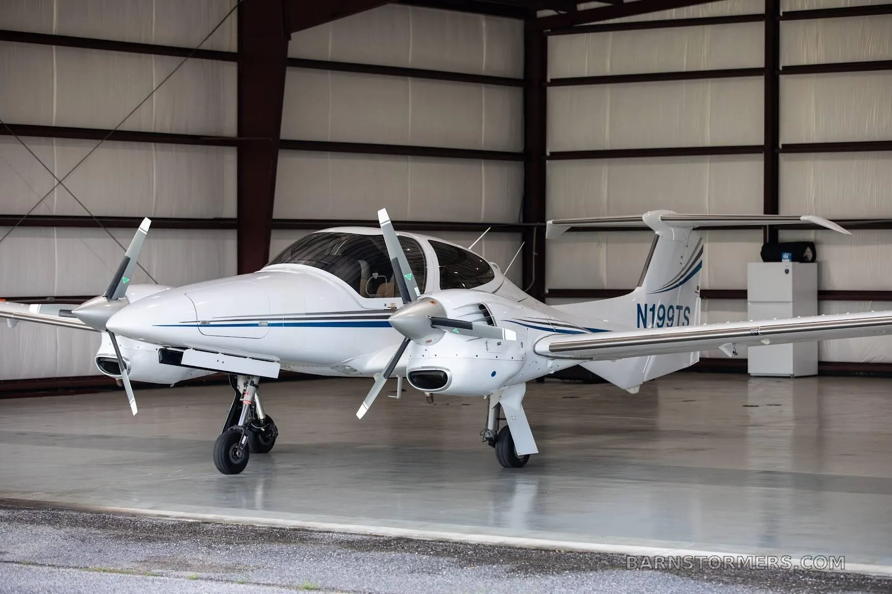 A white Diamond DA42 aircraft with blue stripes, registration N199TS, parked inside a hangar.