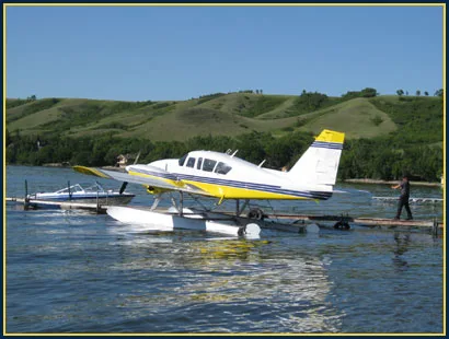 A white and yellow single-engine floatplane is docked on a lake with green hills in the background.