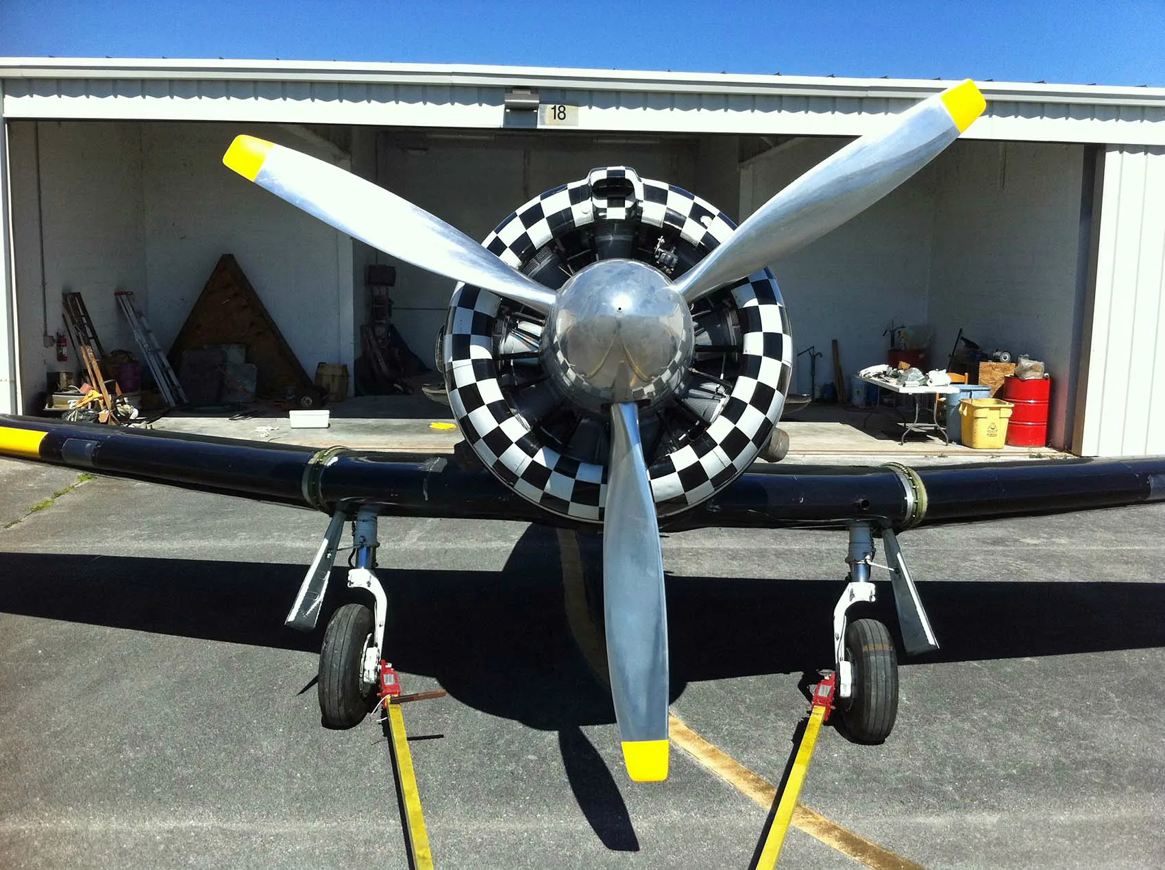 A head-on view of a black warbird aircraft with a large propeller and checkered cowling, parked in front of a hangar.