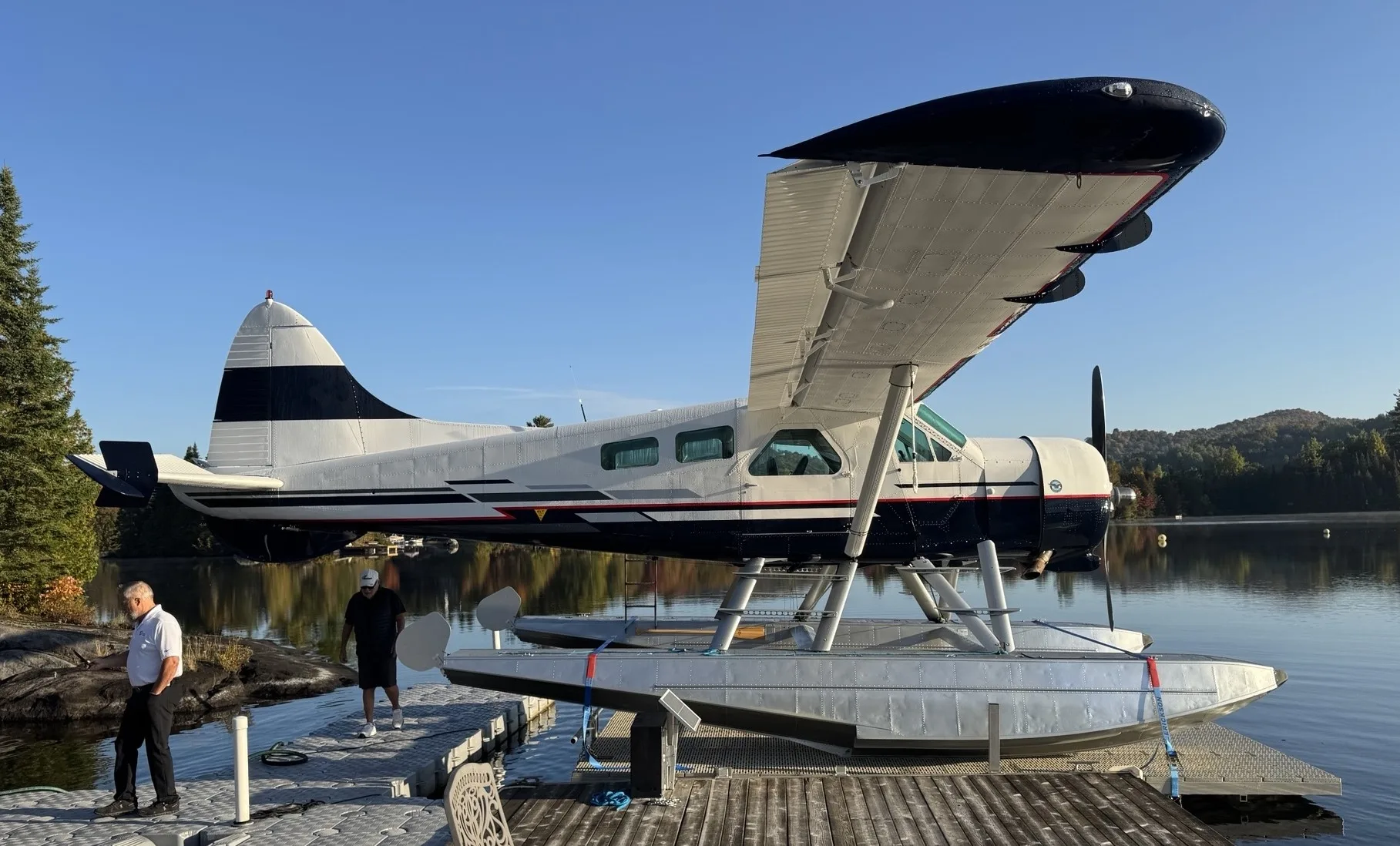 A white and dark blue floatplane with silver floats docked at a pier on a calm lake with trees and hills in the background.