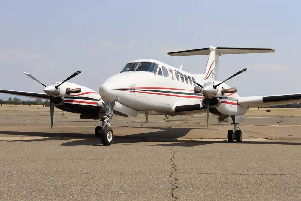 Front view of a white Beechcraft King Air with red and black stripes and propellers on an airport tarmac.