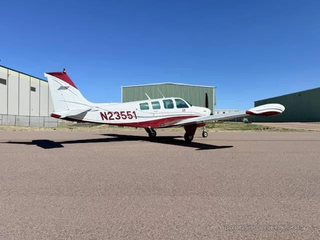 A white and red Beechcraft Bonanza aircraft is parked on an asphalt runway with a hangar in the background.