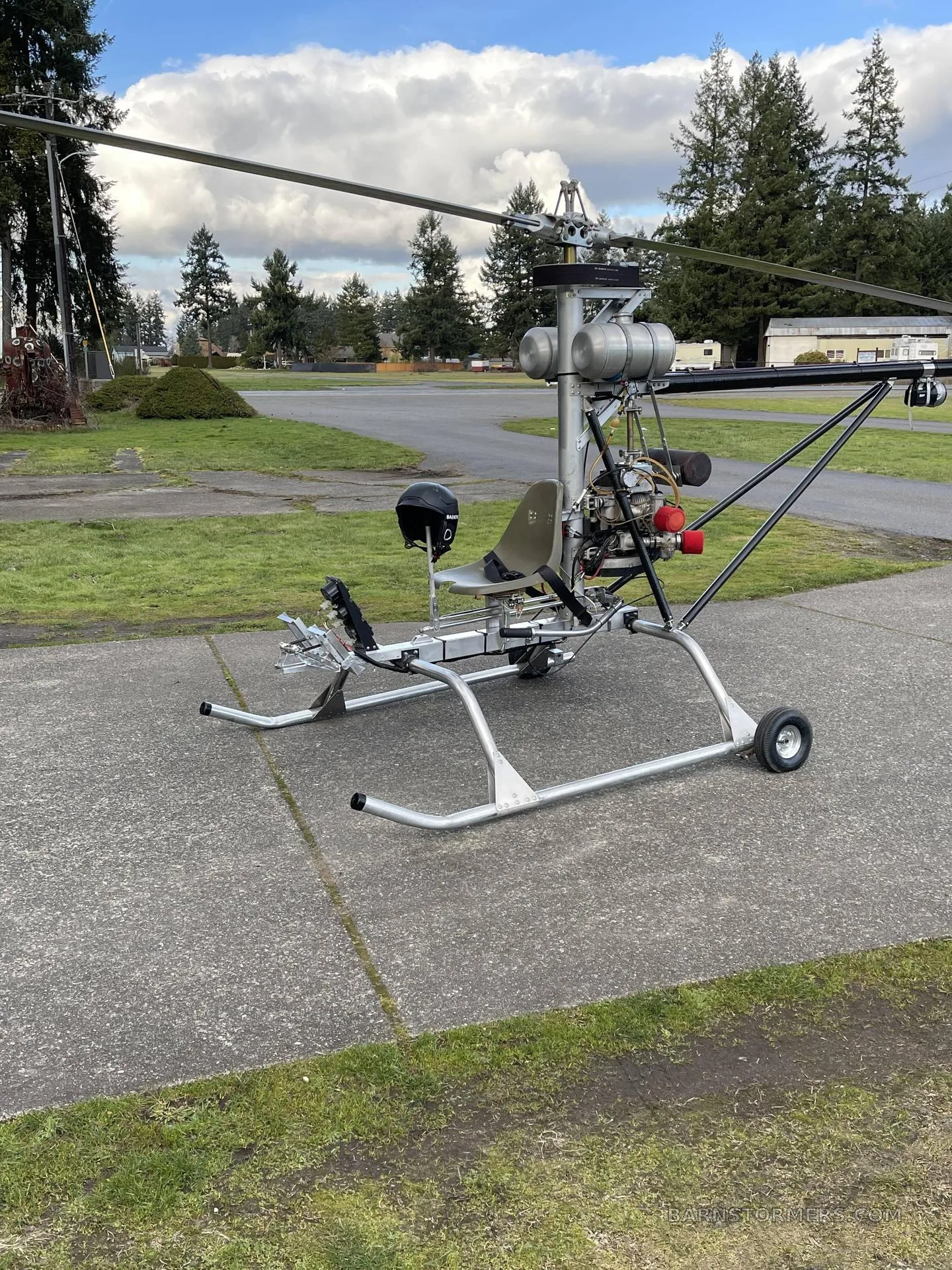 A single-seat ultralight helicopter with an open frame and a black helmet resting on the seat, parked on concrete with trees in the background.