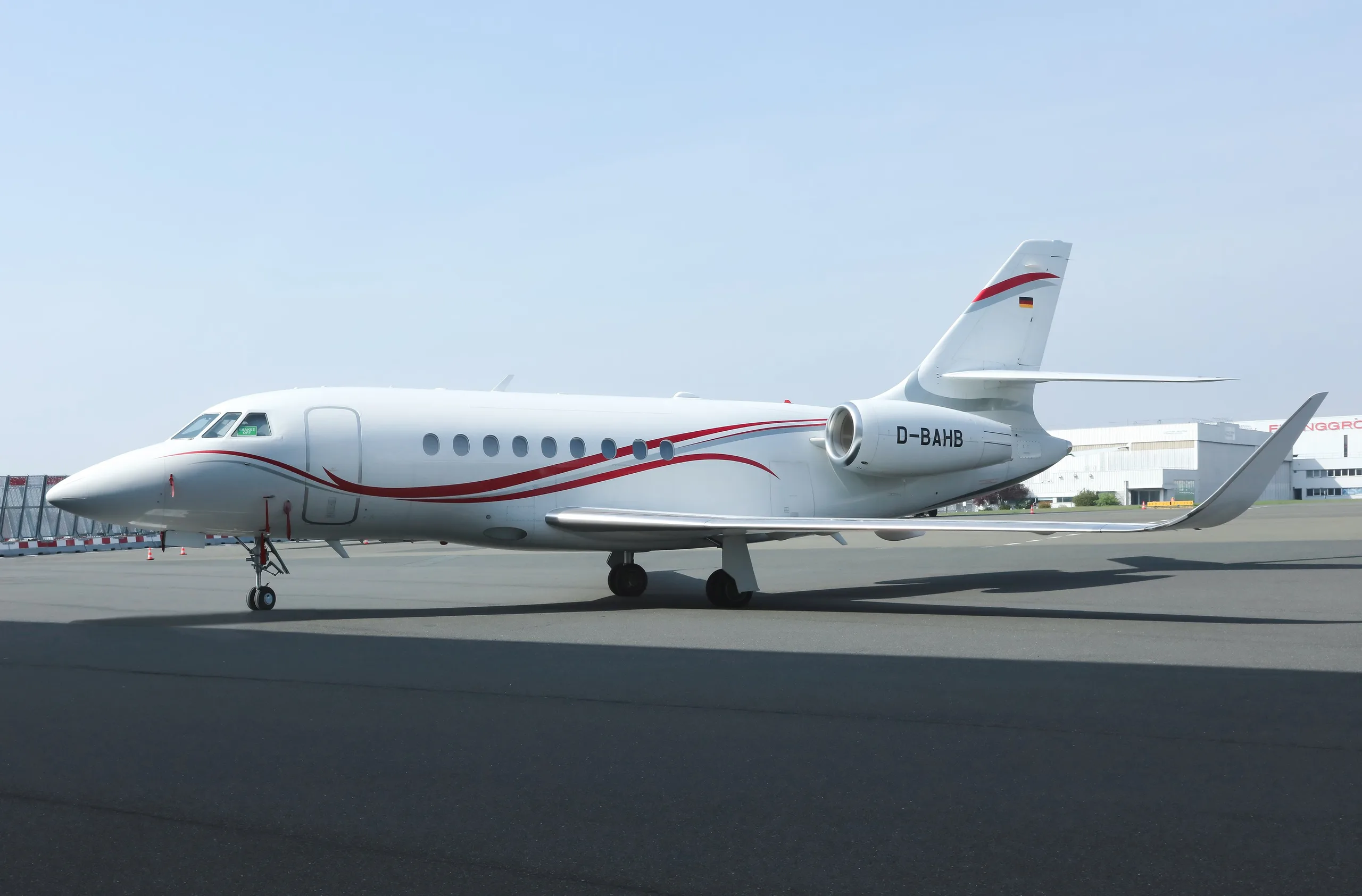 A white business jet with red and grey stripes and German flag on the tail, parked on an airport apron.