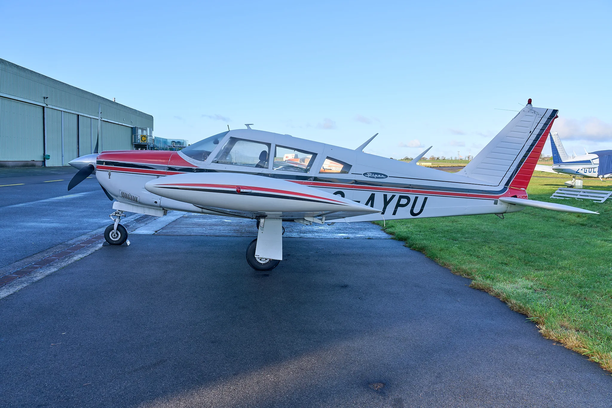 A side view of the white and red Piper Cherokee G-AYPU parked next to a green hangar.