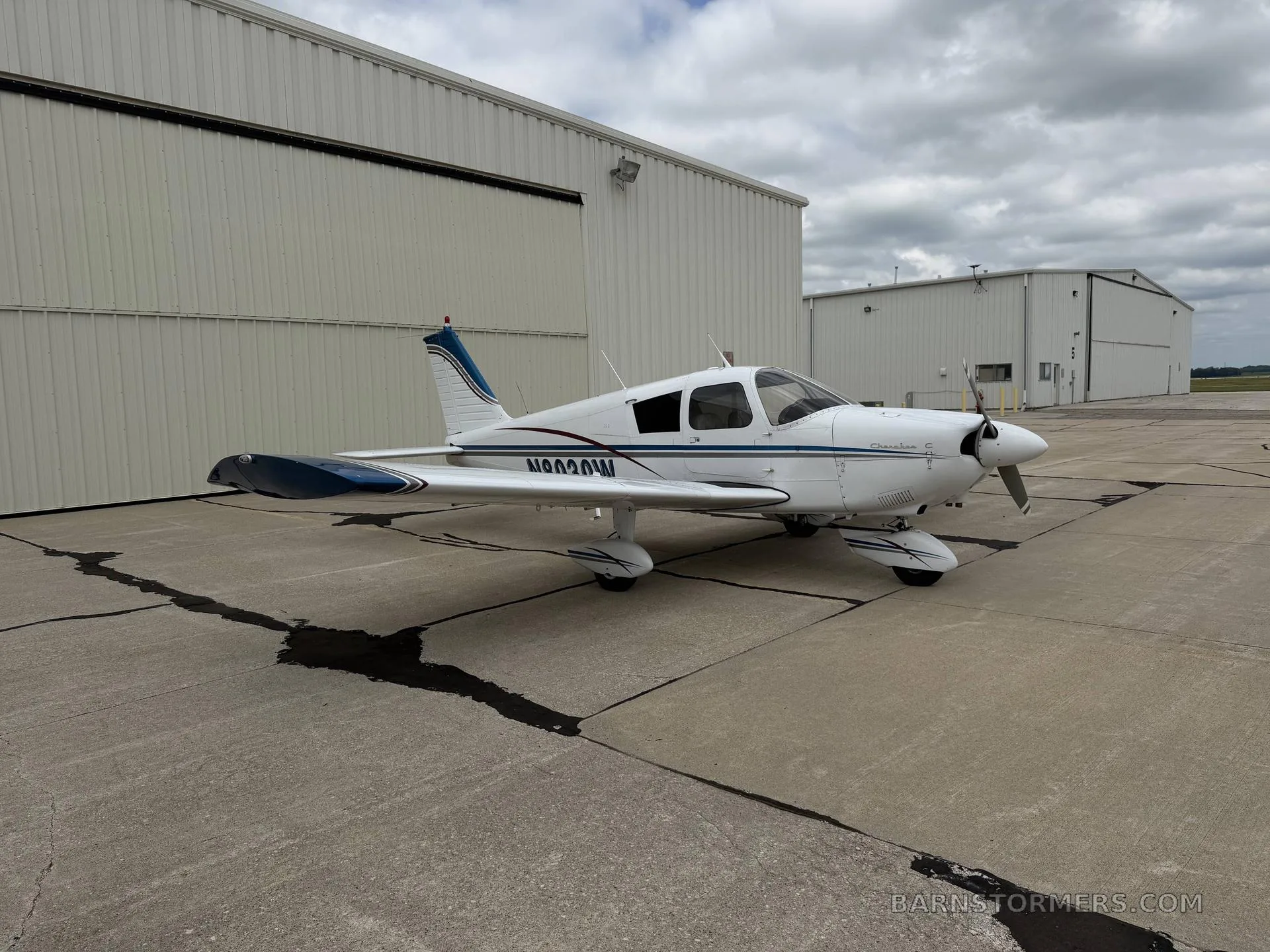 A white Piper Cherokee aircraft with blue accents, viewed from the left side, parked on an asphalt apron next to a hangar.