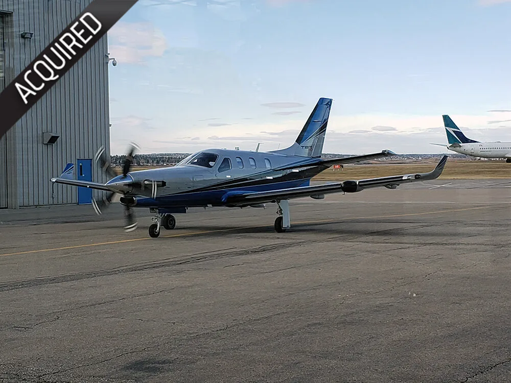 A silver and blue Daher TBM turboprop aircraft is parked on an airport tarmac with a hangar and another plane in the background.