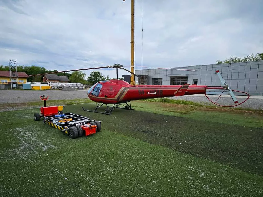 A red helicopter with gold stripes is parked on a green helipad next to a yellow ground handling cart, with a building and crane in the background.
