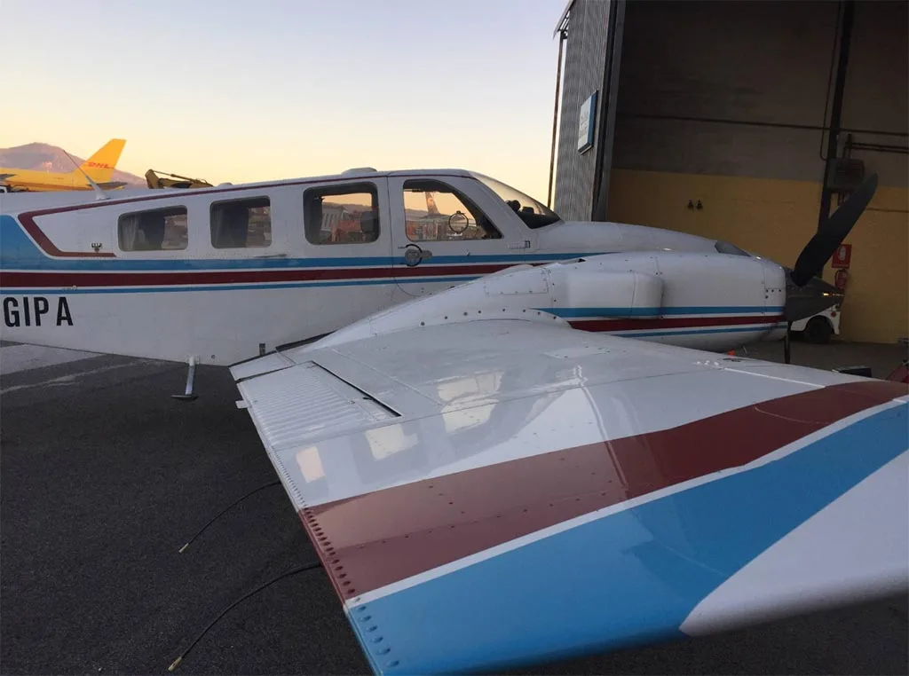 Side view of a white twin-engine aircraft with blue and red stripes, registration I-GIPA, parked next to a hangar at sunset.