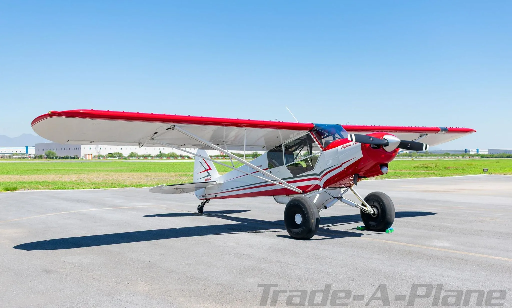 A red and white Super Cub aircraft with large tundra tires parked on an airfield under a clear blue sky.