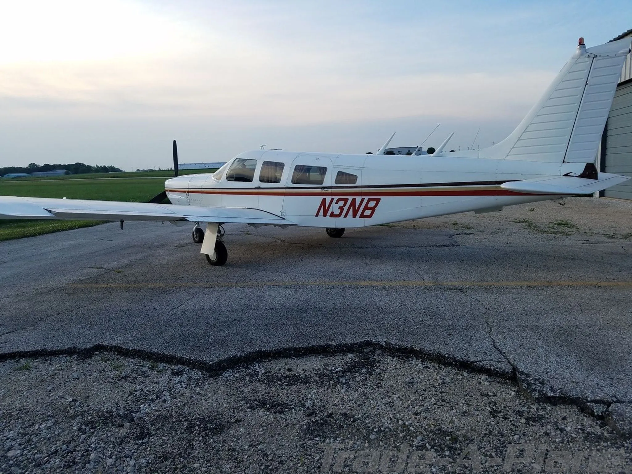 A white Piper aircraft with red and brown stripes, registration N3NB, parked on a cracked asphalt apron.