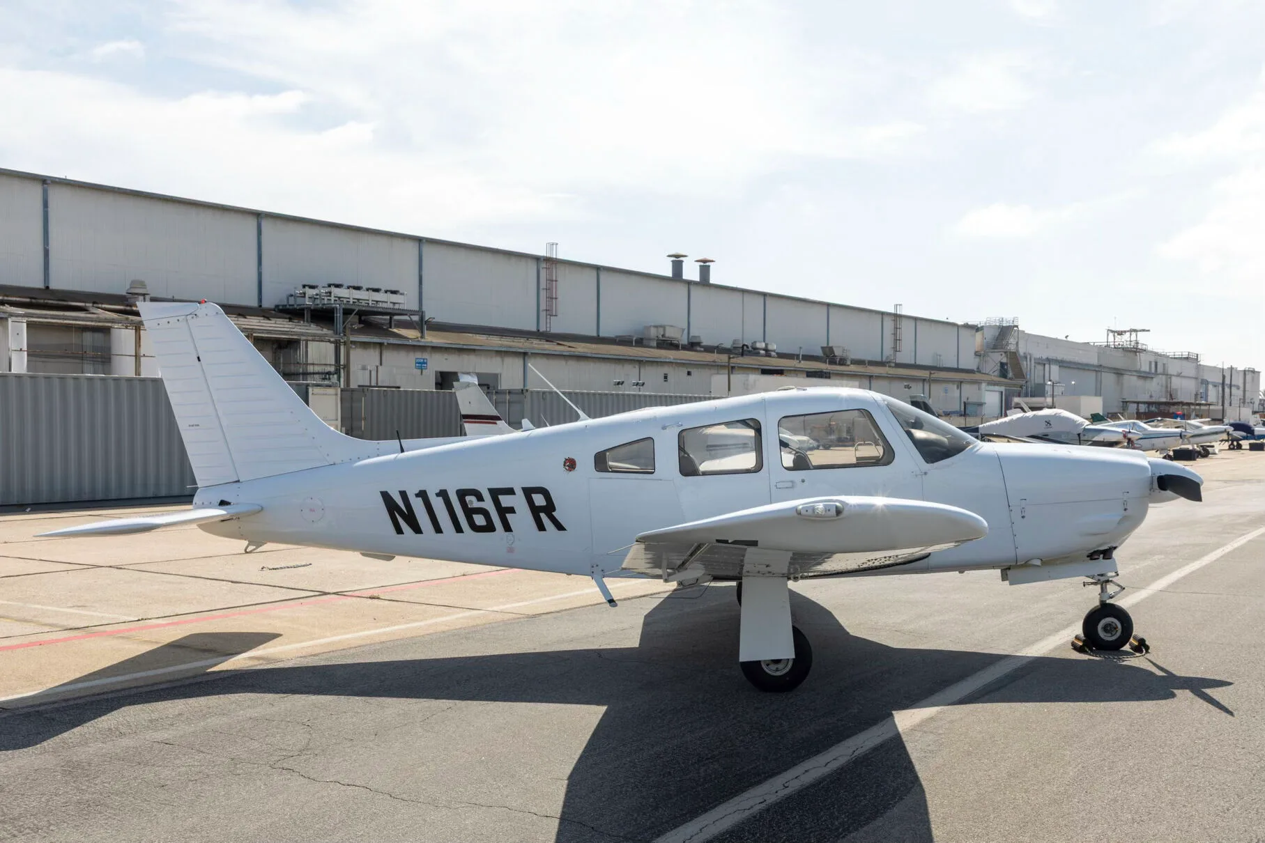 A white Piper PA-28-181 Archer III aircraft, registration N116FR, parked on an airfield in front of hangars.
