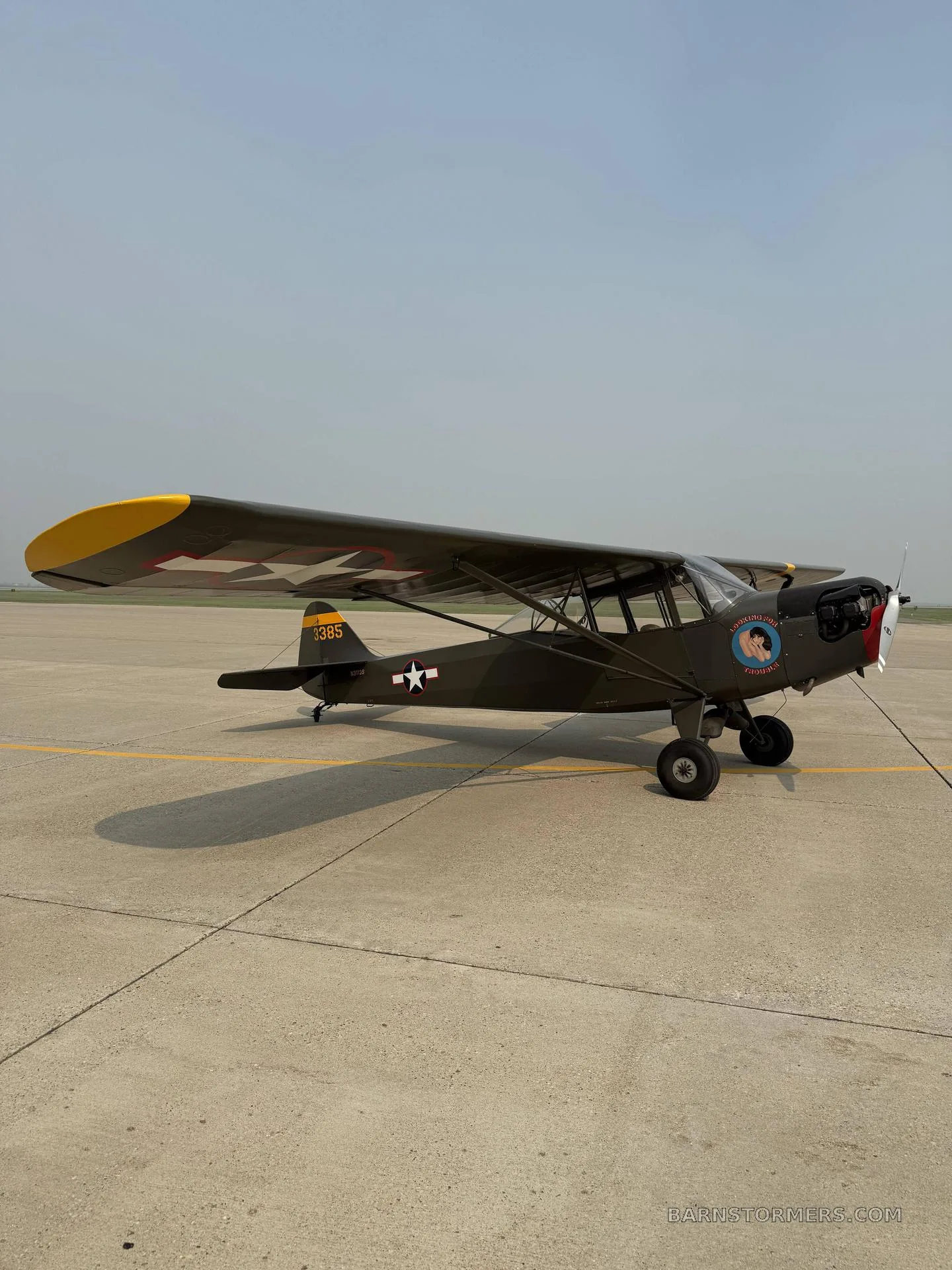 A side view of a military green Piper Cub-like aircraft with yellow wingtips and nose art, parked on an airfield.
