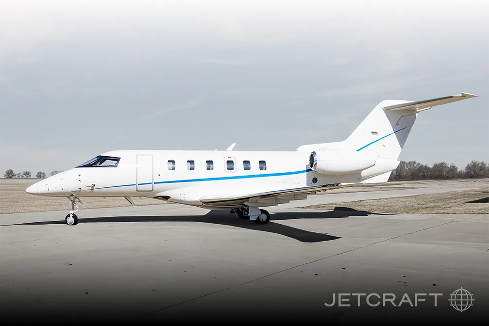 A white business jet with blue stripes is parked on an asphalt tarmac under a clear sky.