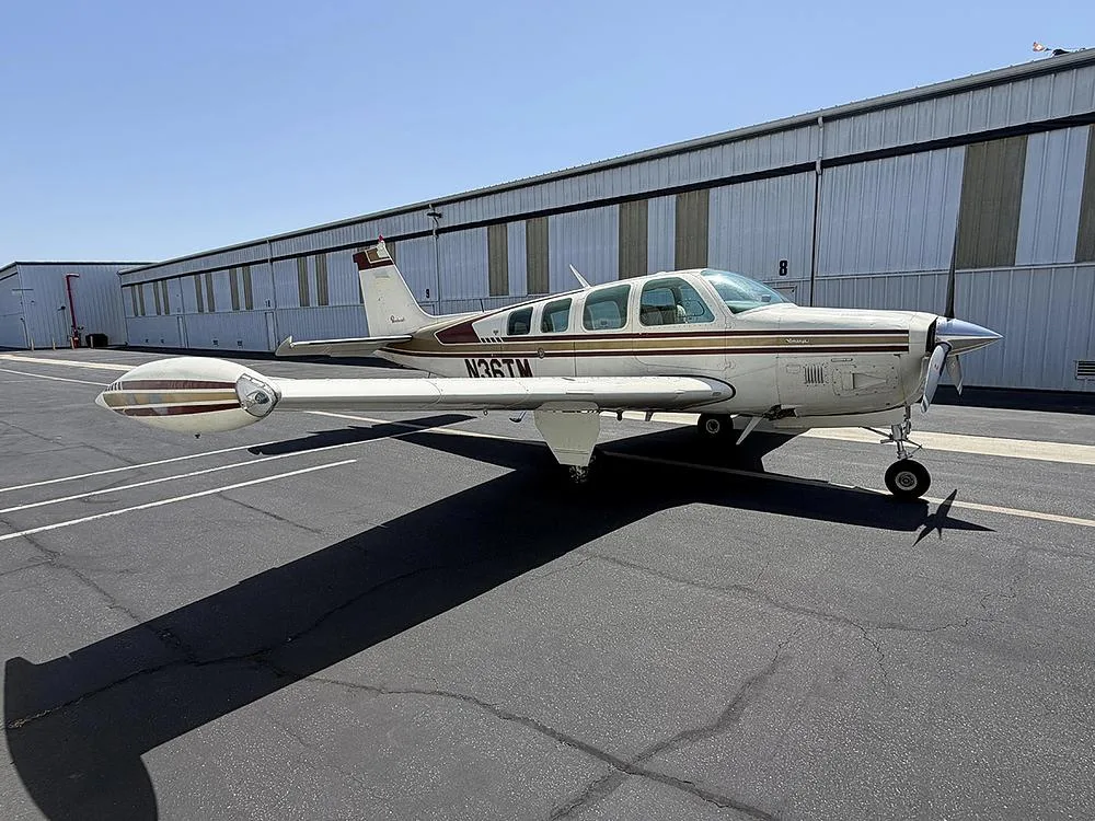 A side view of a white and gold Beechcraft Bonanza N36TM with tip tanks, parked on an asphalt ramp.