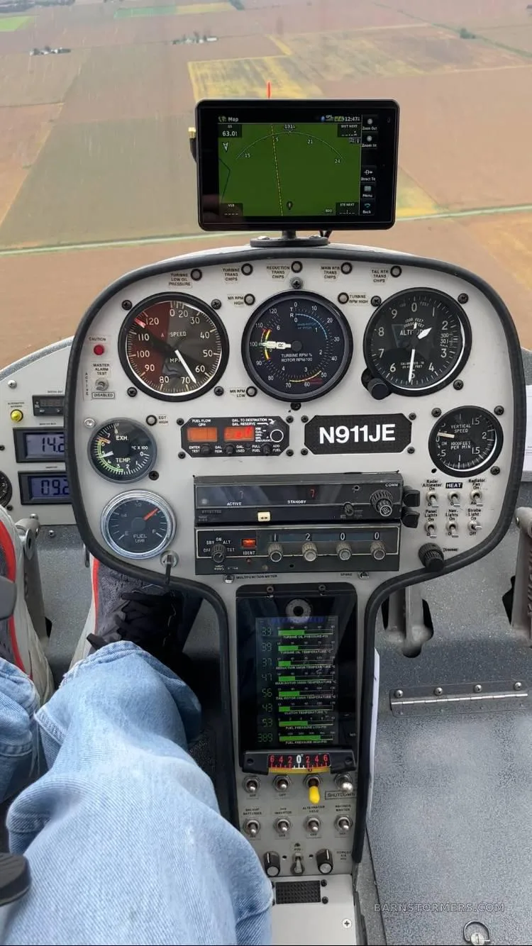 The cockpit of a helicopter (N911JE) in flight, showing the instrument panel, GPS display, and pilot's legs over agricultural fields.