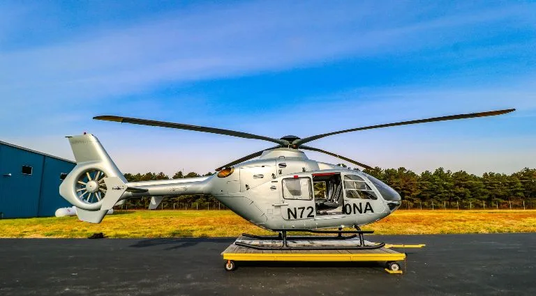 A silver helicopter, registration N72ONA, sits on a yellow dolly on an asphalt pad next to a blue hangar under a blue sky.