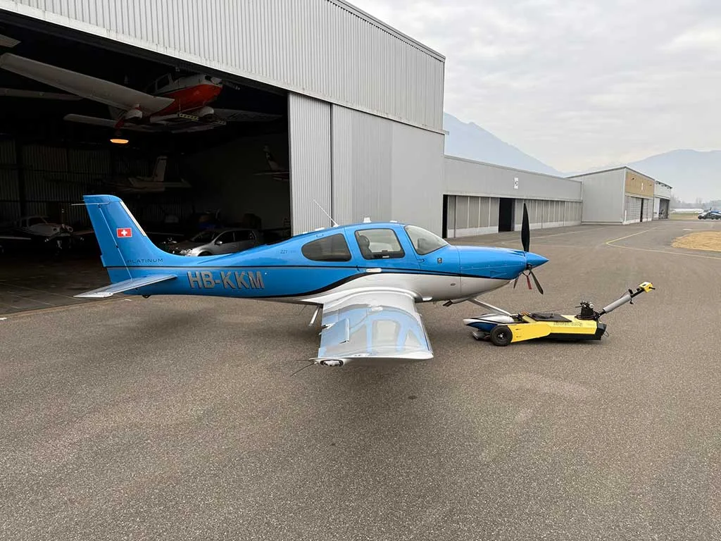 A side view of a blue and silver Cirrus SR22T aircraft with registration HB-KKM parked outside a hangar.