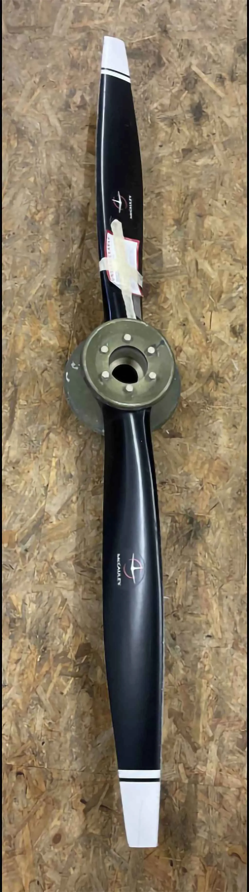 A black McCauley aircraft propeller with white tips and a red label is shown against a wooden background.