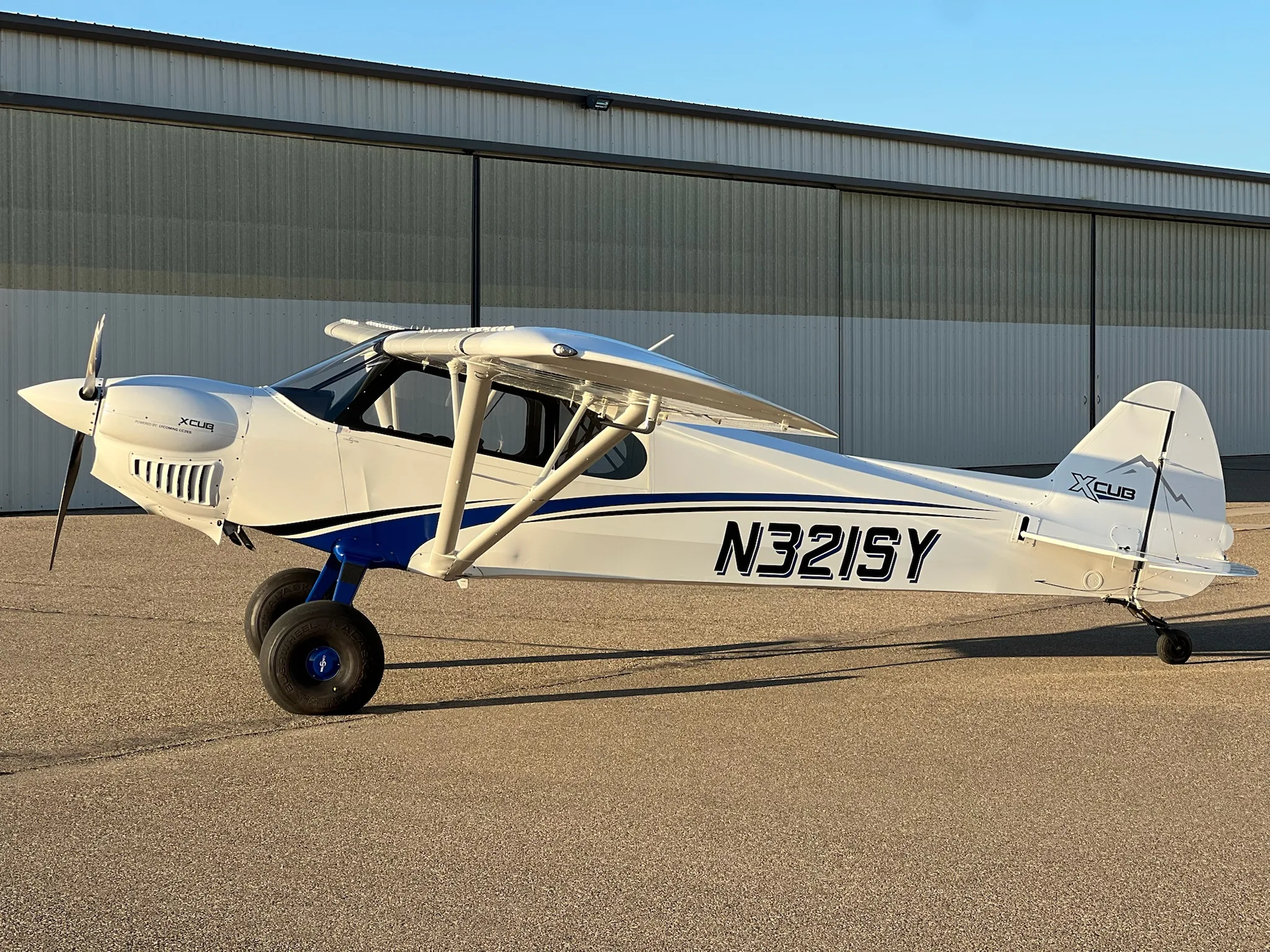 A white X-Cub aircraft with blue stripes and registration N321SY parked on an airfield in front of a hangar.