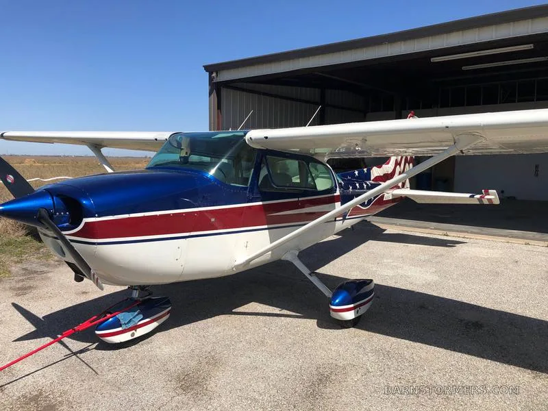 A front-quarter view of a Cessna 172 with an American flag paint scheme, parked outside a hangar.