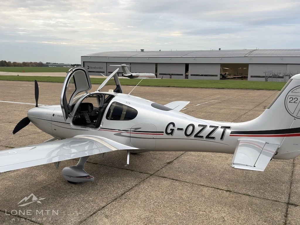 A silver Cirrus SR22T aircraft with registration G-OZZT and open door, parked on an airfield tarmac.