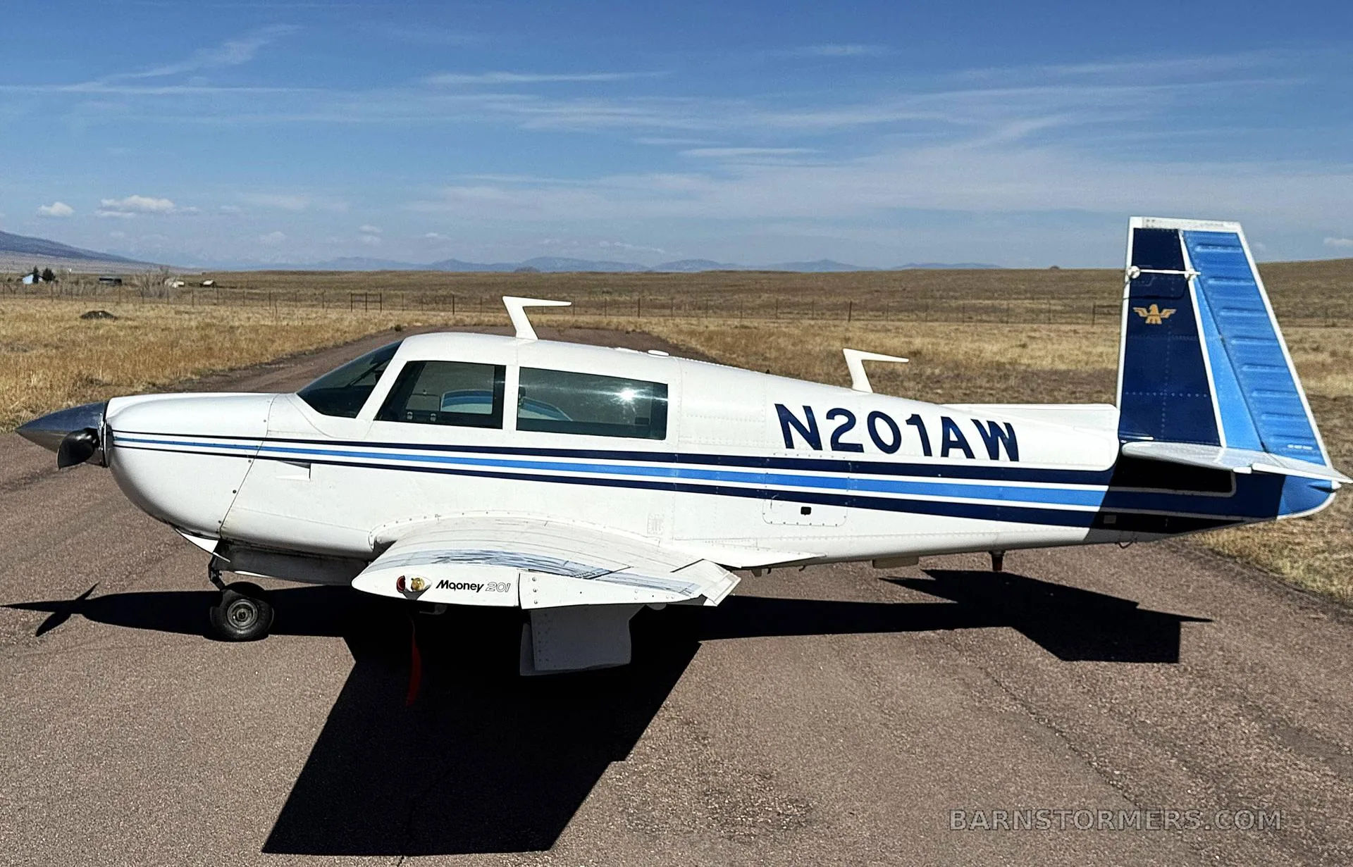 A white Mooney M20J aircraft with blue stripes and tail, registration N201AW, parked on an asphalt runway with mountains in the background.