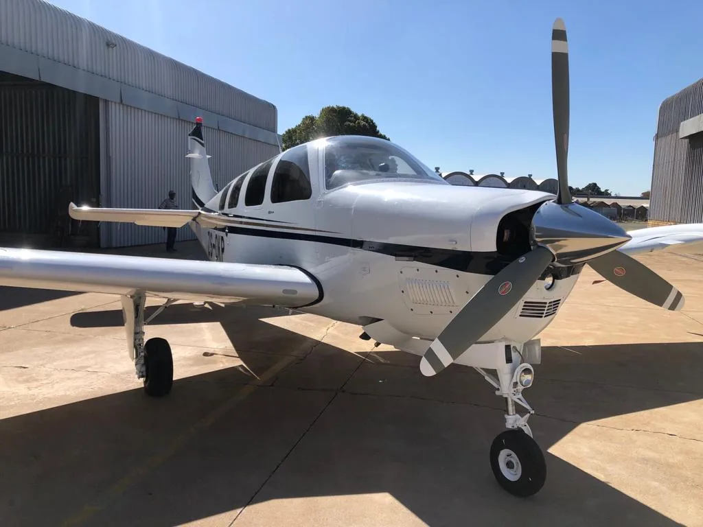 A white single-engine propeller aircraft with a dark stripe, parked on an apron in front of a metal hangar under a clear sky.