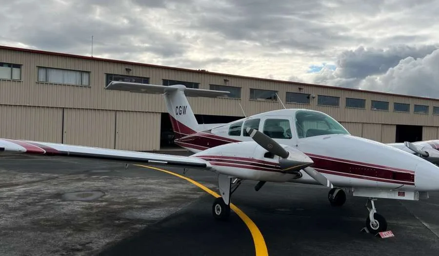 A white twin-engine propeller aircraft with maroon stripes is parked on an tarmac in front of a hangar under a cloudy sky.