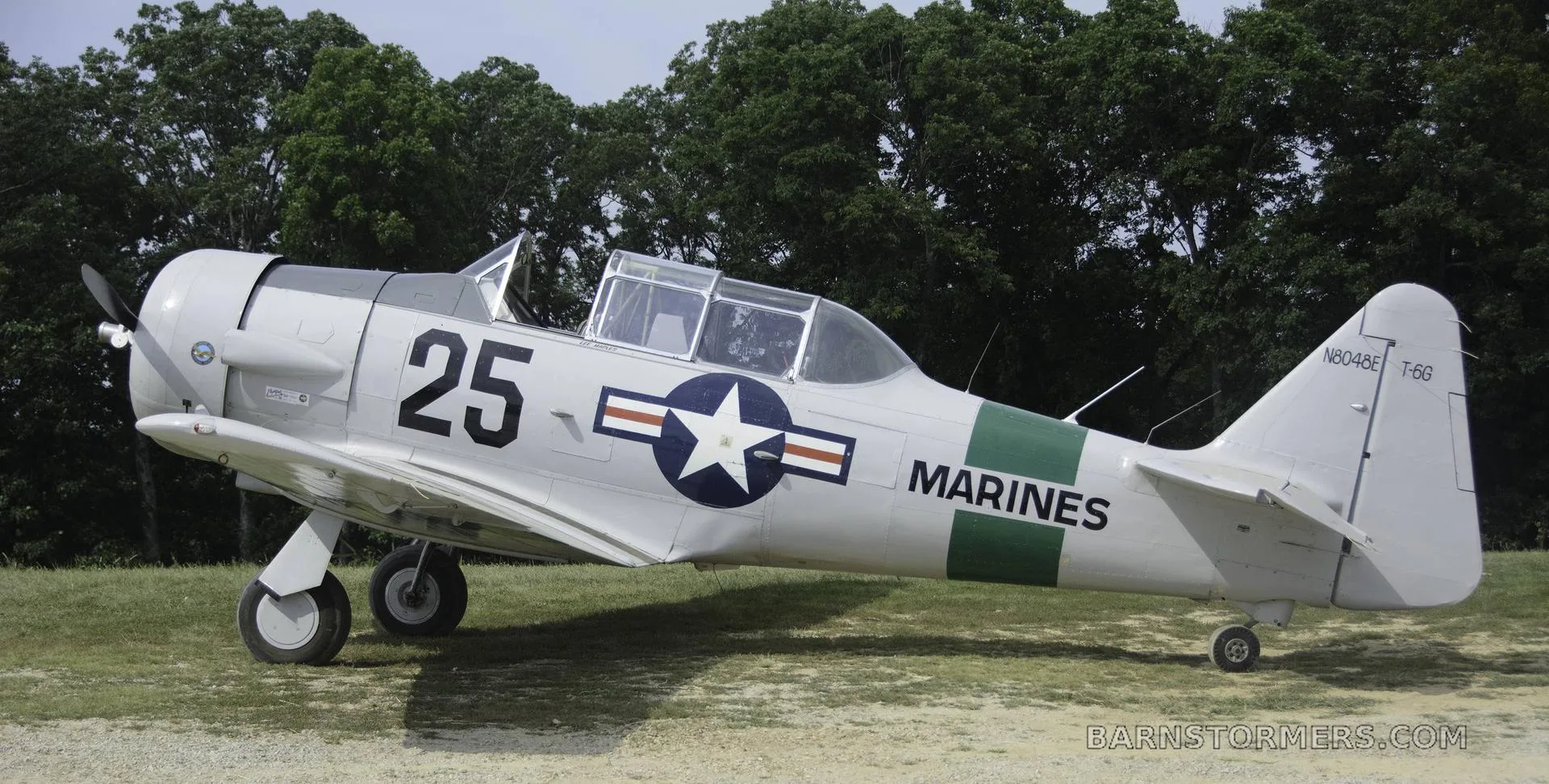 A light gray T-6G Texan aircraft with "MARINES" and "25" markings, a US star insignia, and a green stripe, parked on a grassy area.