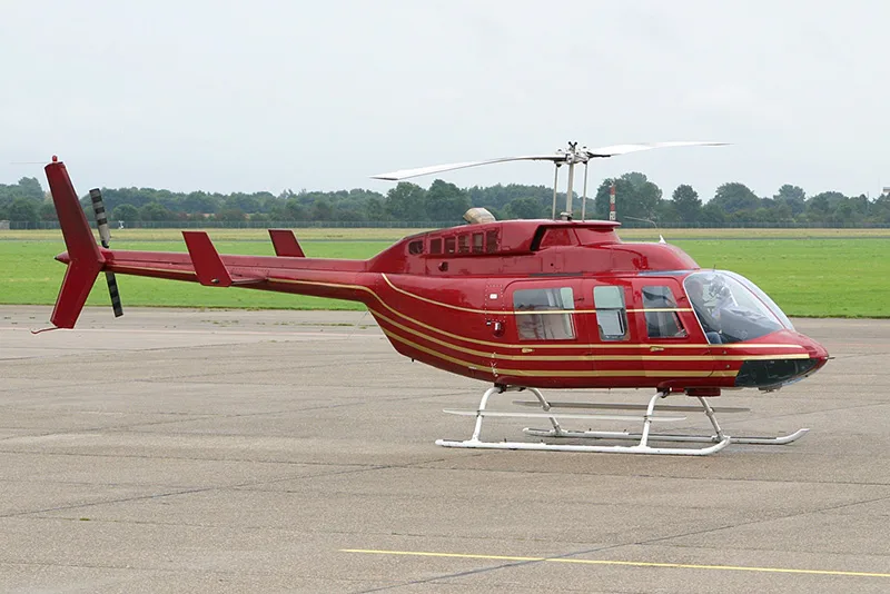 A red Bell 206 helicopter with gold stripes sits on the tarmac.